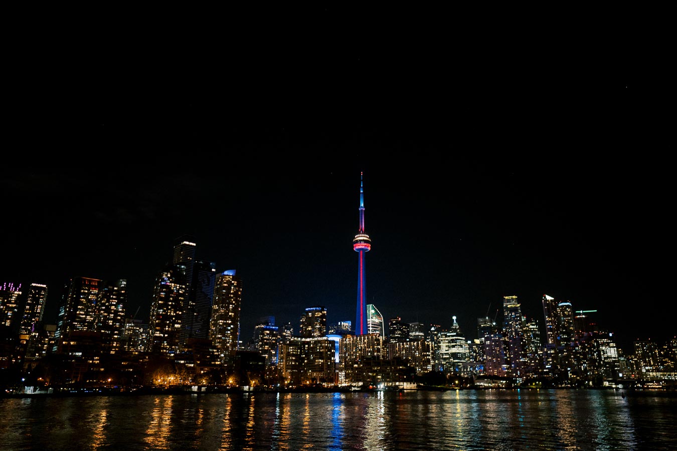 Night skyline of Toronto featuring the illuminated CN Tower with city lights reflecting on Lake Ontario.