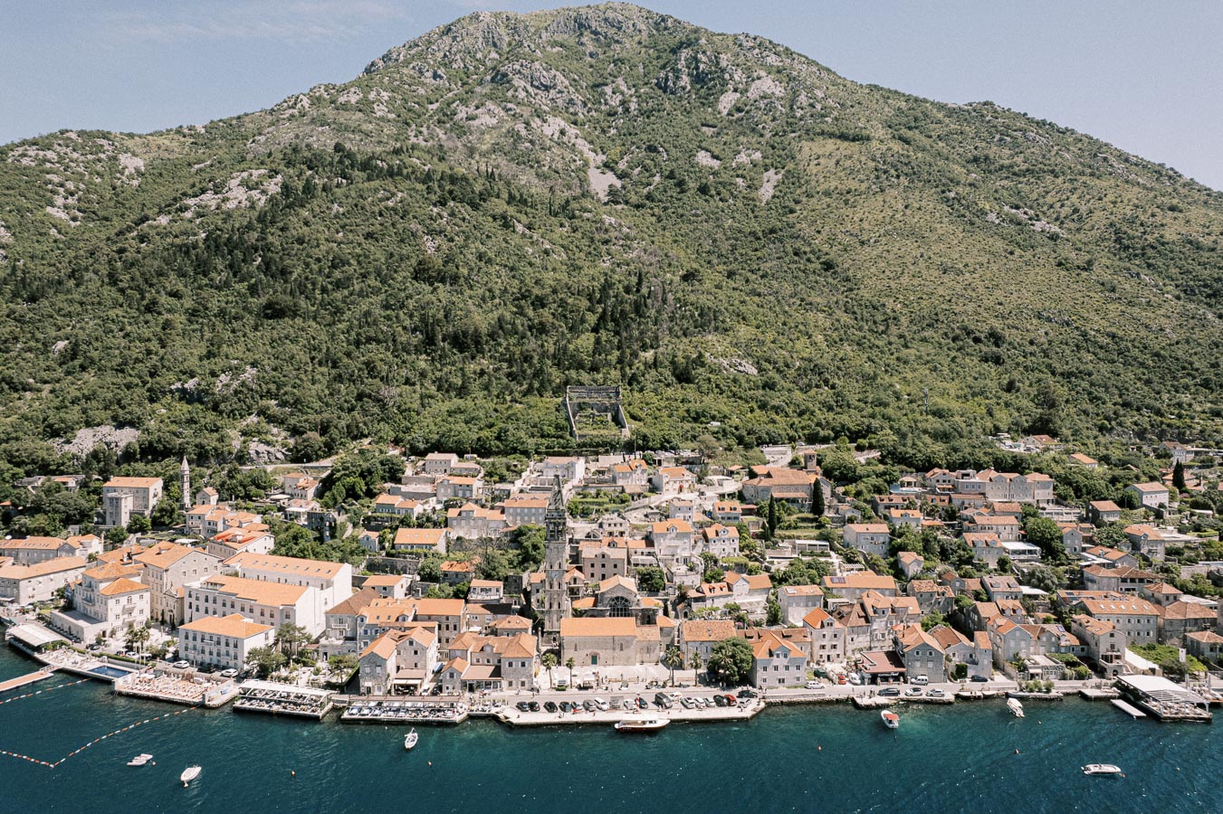 Aerial view of a picturesque coastal town with terracotta-roofed buildings nestled along a clear blue waterfront, set against a backdrop of lush green hills.
