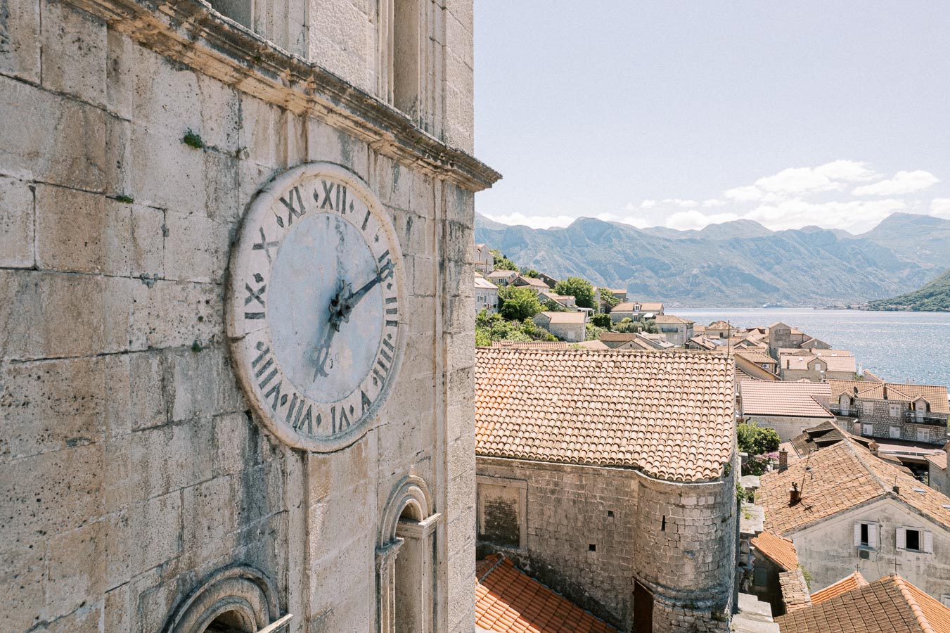 Historic stone clock tower overlooking a coastal village with terracotta rooftops and scenic mountains by the sea on a clear day.