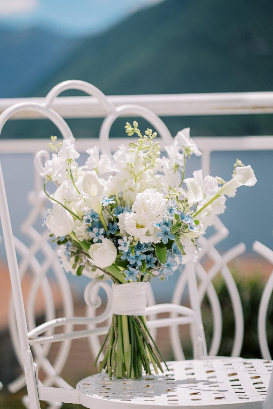 A beautiful bouquet of white and blue flowers on a vintage white chair, set against a serene lake and mountain backdrop.