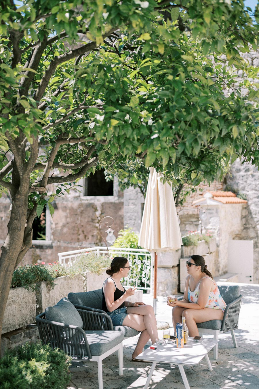 Two women sitting on outdoor patio furniture in a shaded garden, engaging in conversation with drinks on the table. Stone walls and lush greenery create a serene and relaxing atmosphere.