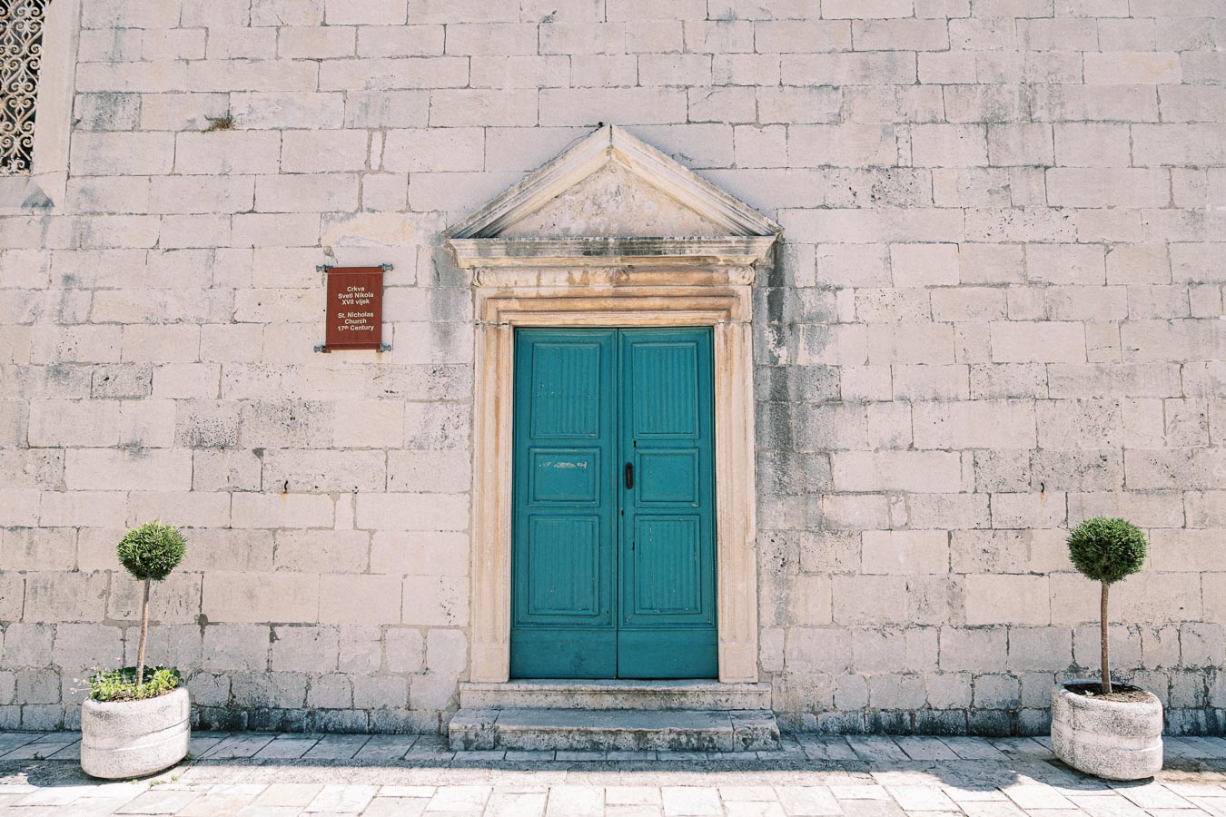 Historic blue wooden door with stone frame on a vintage brick wall, flanked by two potted plants, featuring a plaque for St. Nicholas 17th Century architecture.