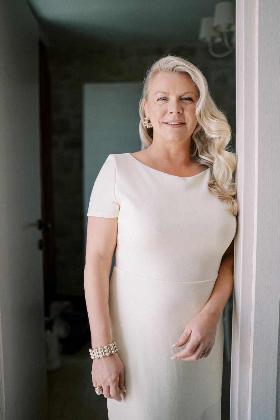 A woman with long, wavy blonde hair wearing a white dress, standing in a doorway and smiling at the camera.