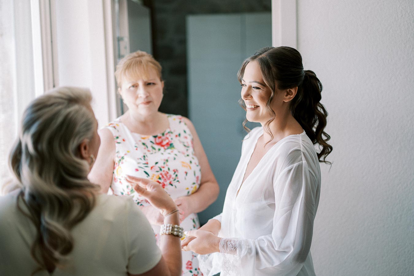 A bride in a white robe smiling while talking to two women, one wearing a floral dress, in a softly lit room near a window.