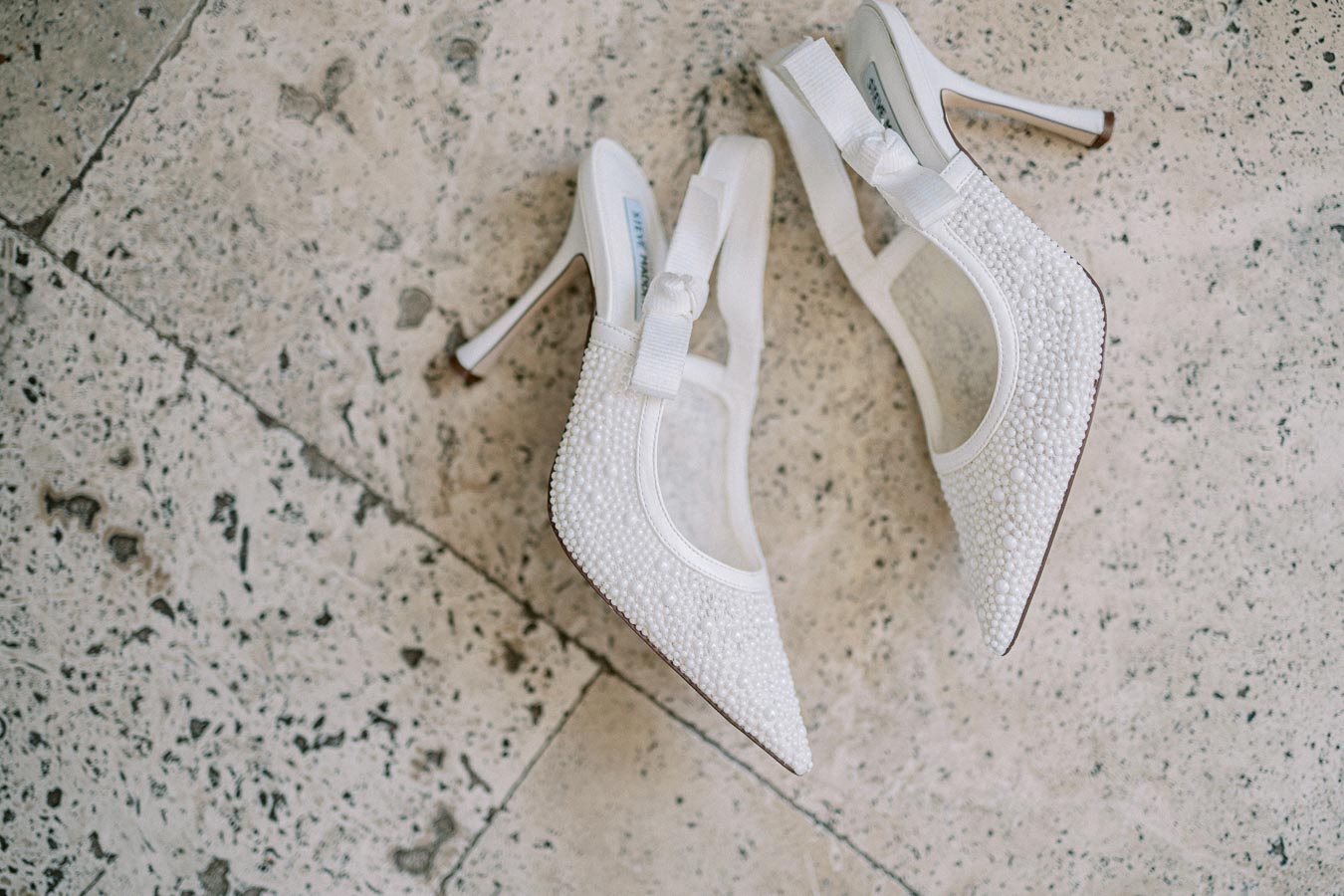 Elegant pair of white high-heeled shoes with intricate bead detailing on a textured marble floor, perfect for weddings or formal occasions.