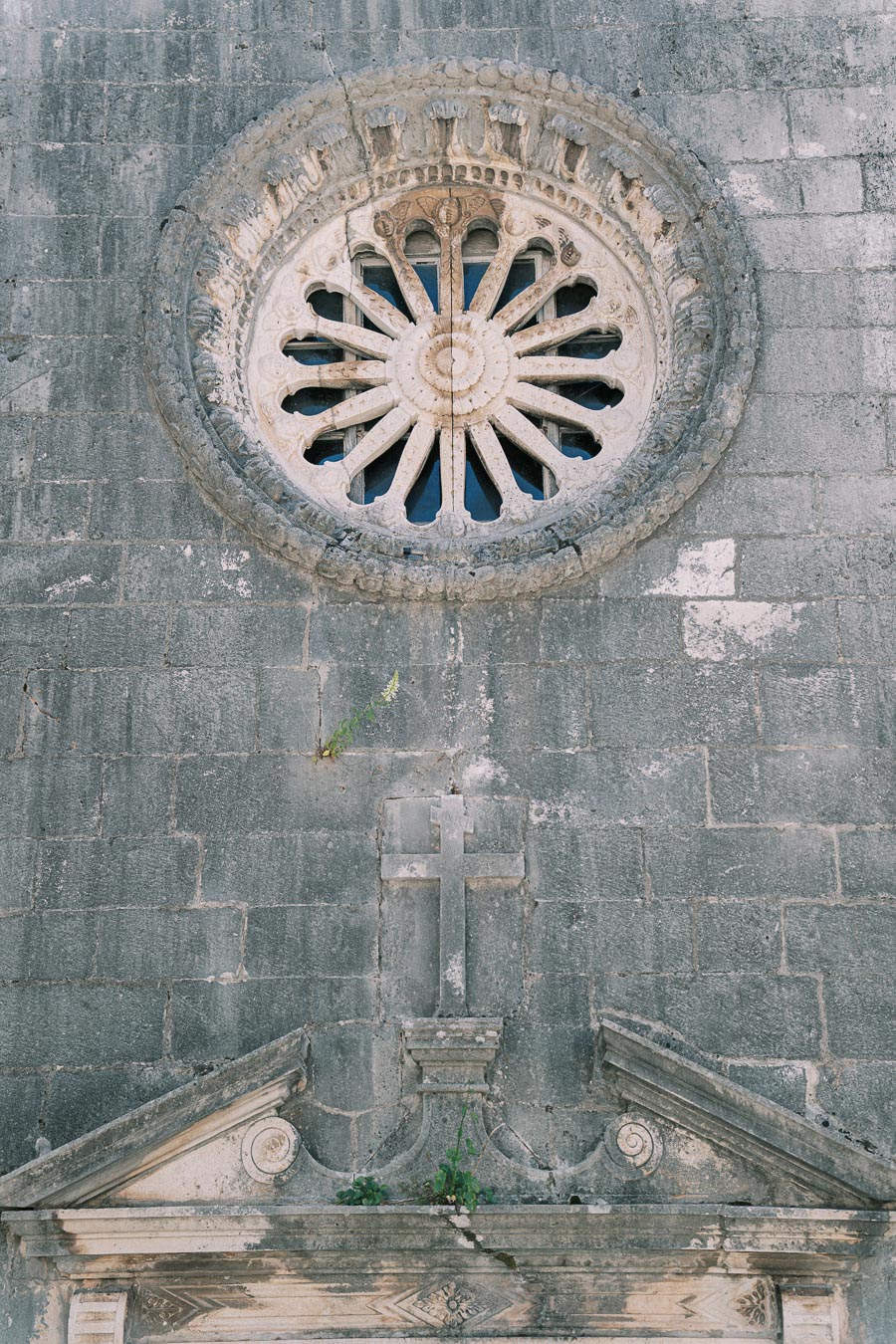 Ancient stone church facade showcasing a detailed circular rose window above an intricately carved cross and pediment, highlighting historic architectural elements.