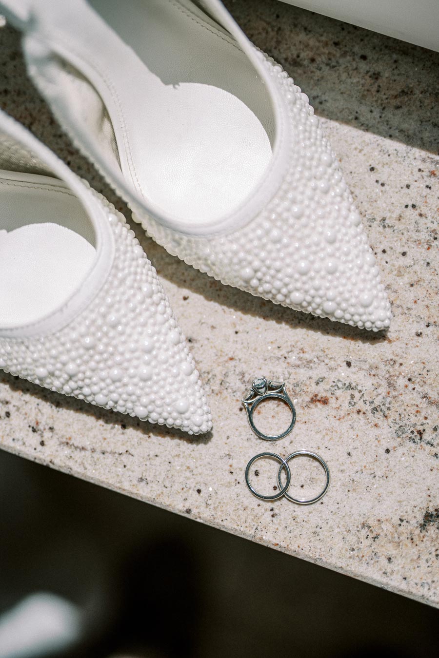 Elegant bridal scene with pearl-embellished white high heels and diamond engagement ring alongside wedding bands on a textured stone surface.