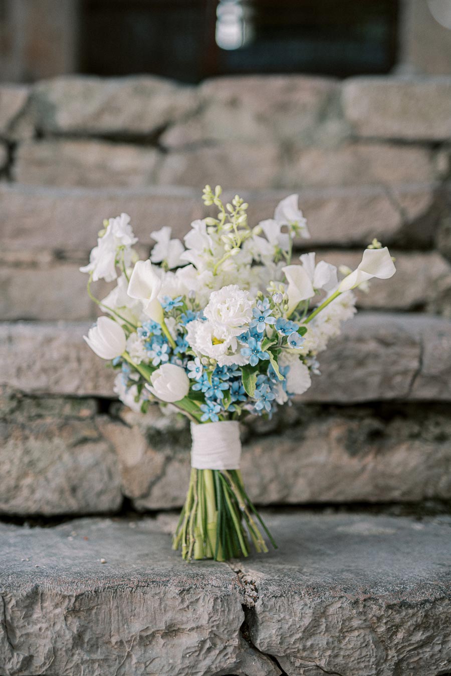 Bridal bouquet with white and blue flowers arranged elegantly against stone steps backdrop.