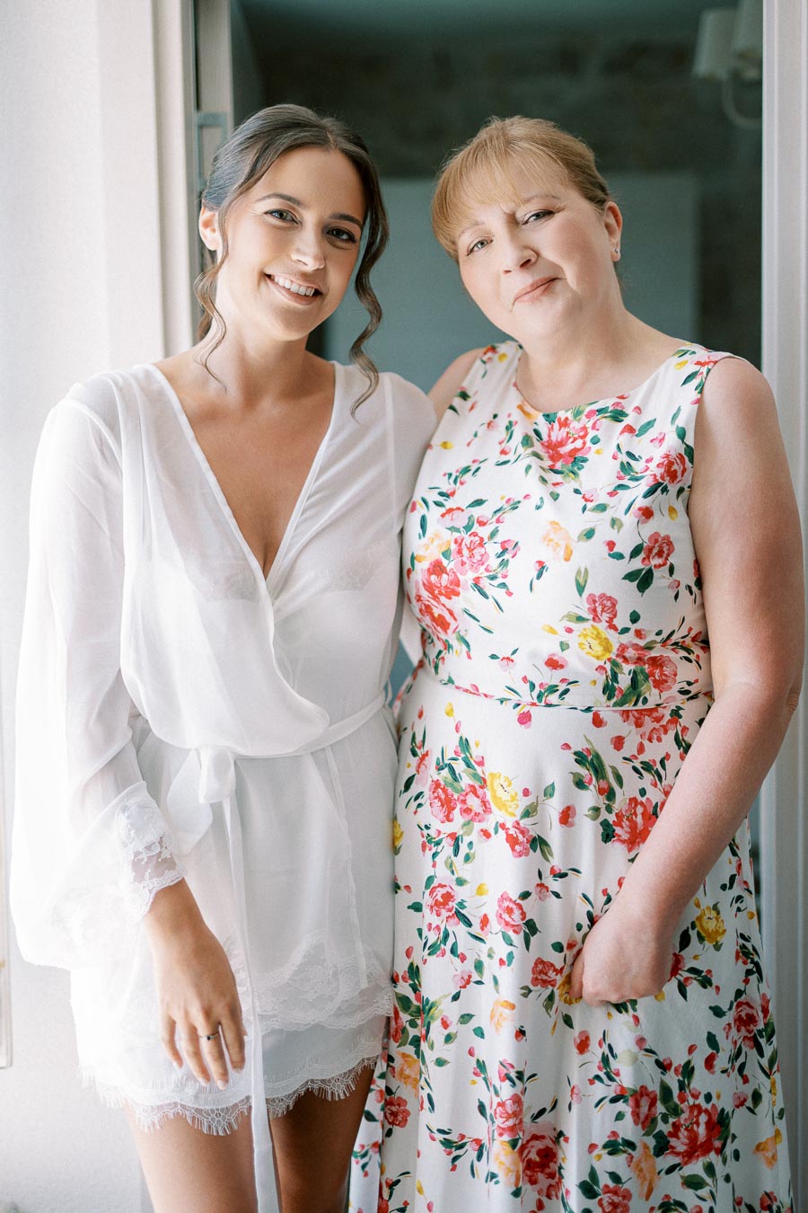 Two women smiling and standing together indoors, one in a white robe and the other in a floral dress.
