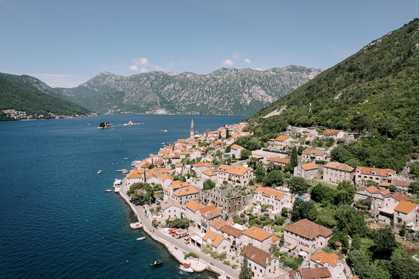 A picturesque coastal village with terracotta-roofed buildings and a church tower, nestled beside a tranquil bay with mountainous terrain in the background under a clear blue sky.