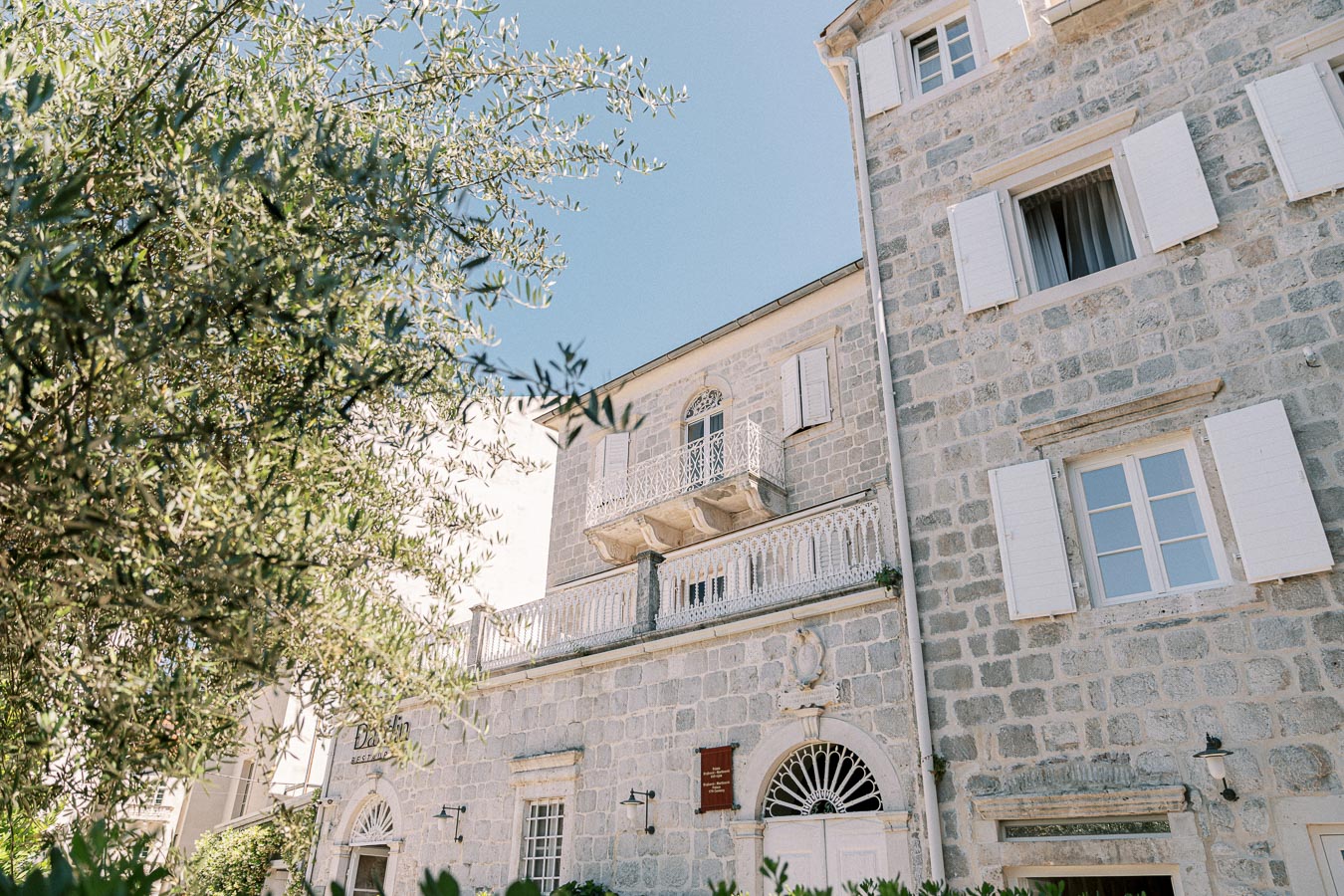 Old stone building with white shutters and ornate balcony against a clear blue sky, surrounded by lush greenery.