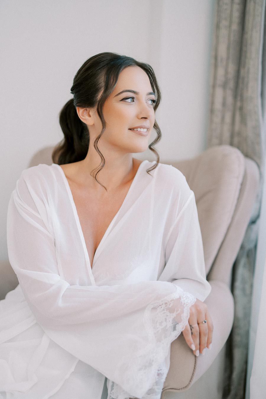 Elegant woman in a white robe with lace details, sitting on a plush chair, gazing thoughtfully out of a window