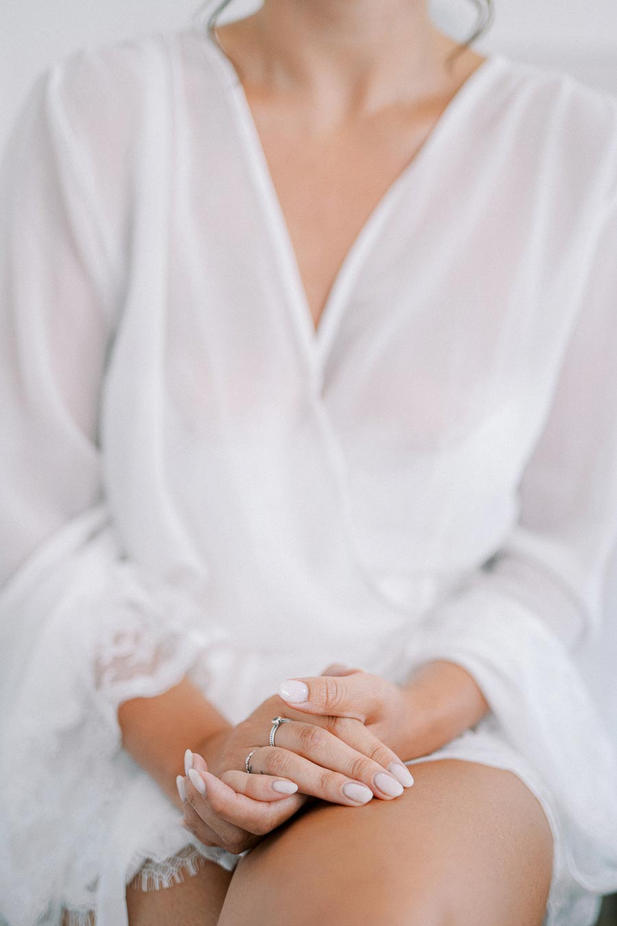 Close-up of a woman in a white robe with delicate lace details, showcasing elegant hands resting on her knee with neatly manicured nails and subtle rings.