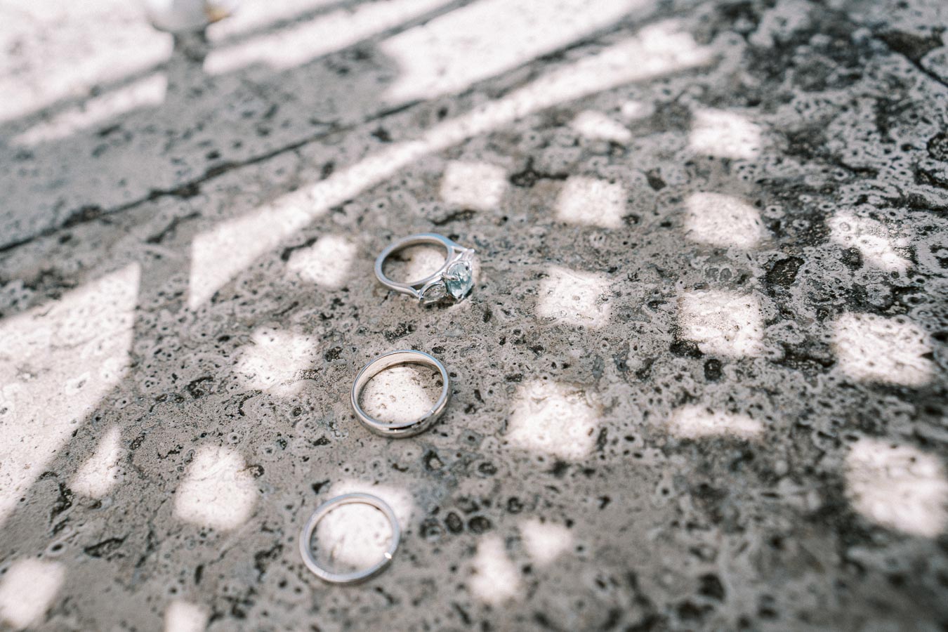 Three wedding rings, including a diamond engagement ring, placed on a textured stone surface with shadow patterns filtering through.