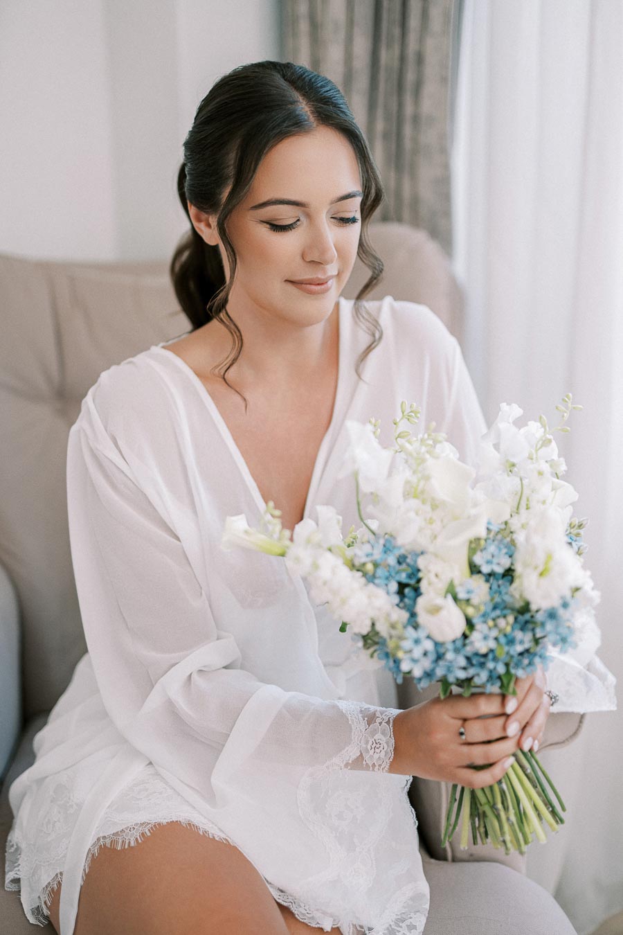 Young woman in a white robe holding a bouquet of blue and white flowers, sitting on a sofa with natural light coming through a window.