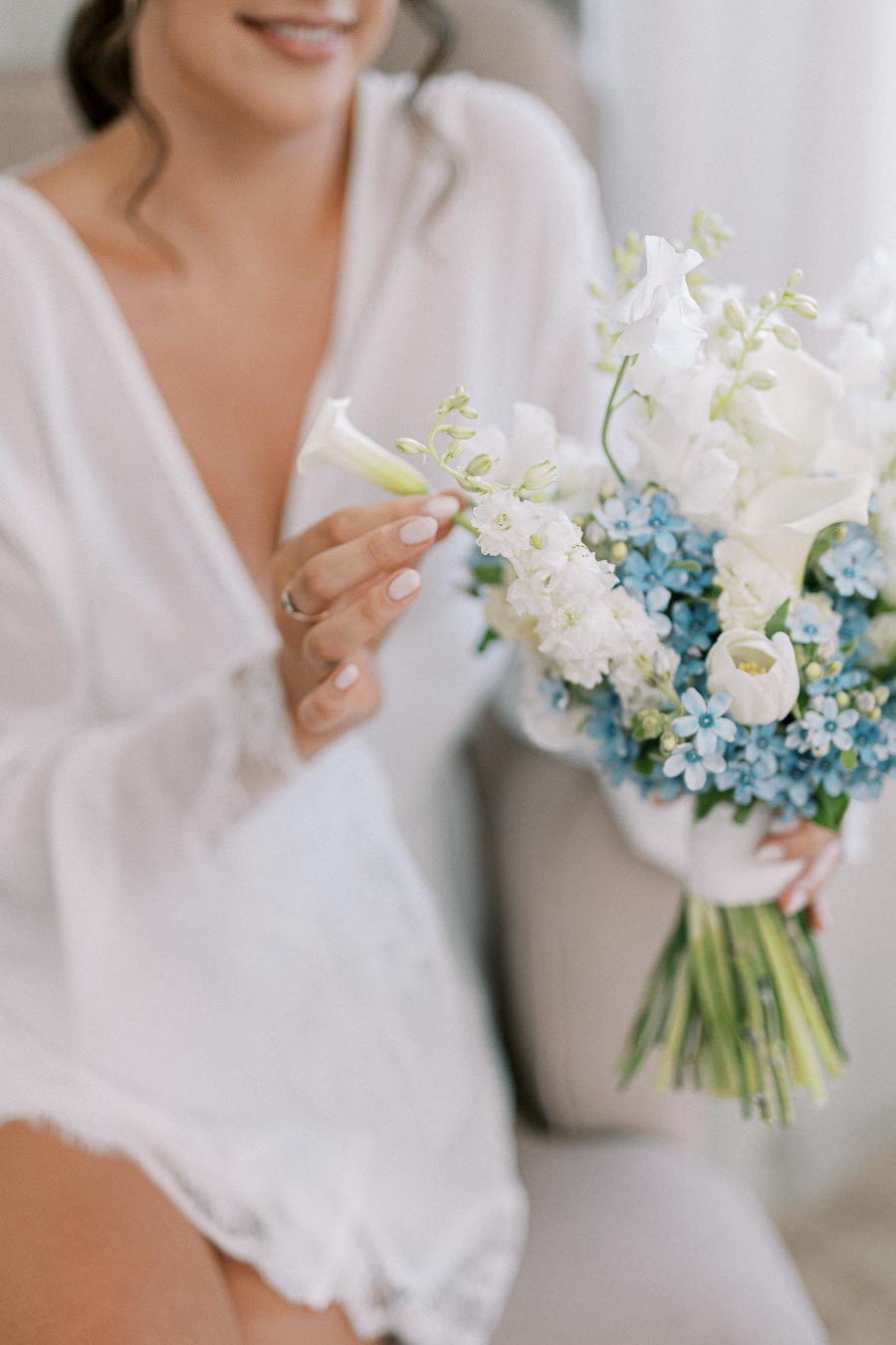 A bride in a white robe holding a bouquet of white and blue flowers, gently touching a bloom.