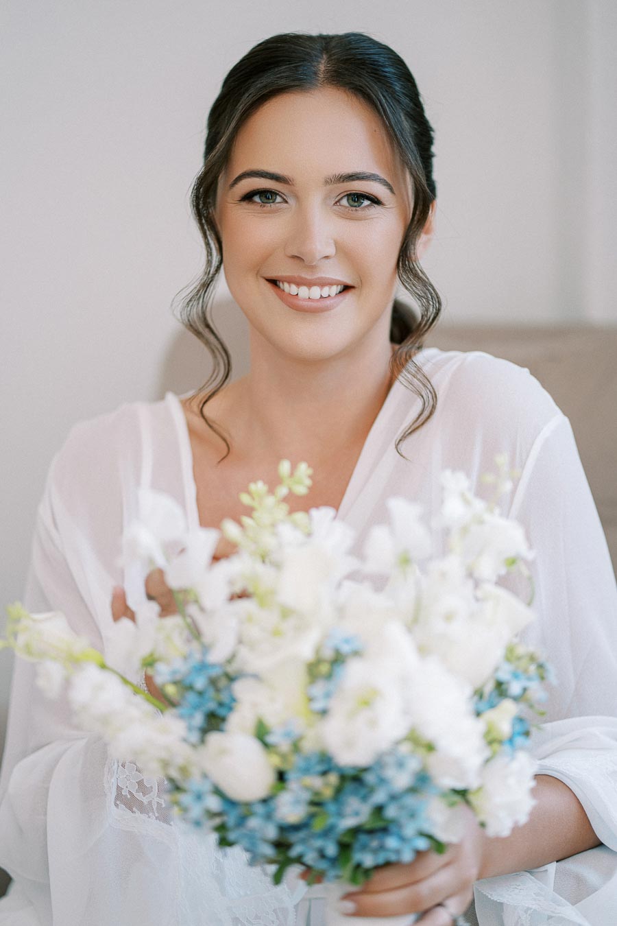 Smiling bride holding a bouquet of blue and white flowers, wearing a white robe with lace details, preparing for wedding day.
