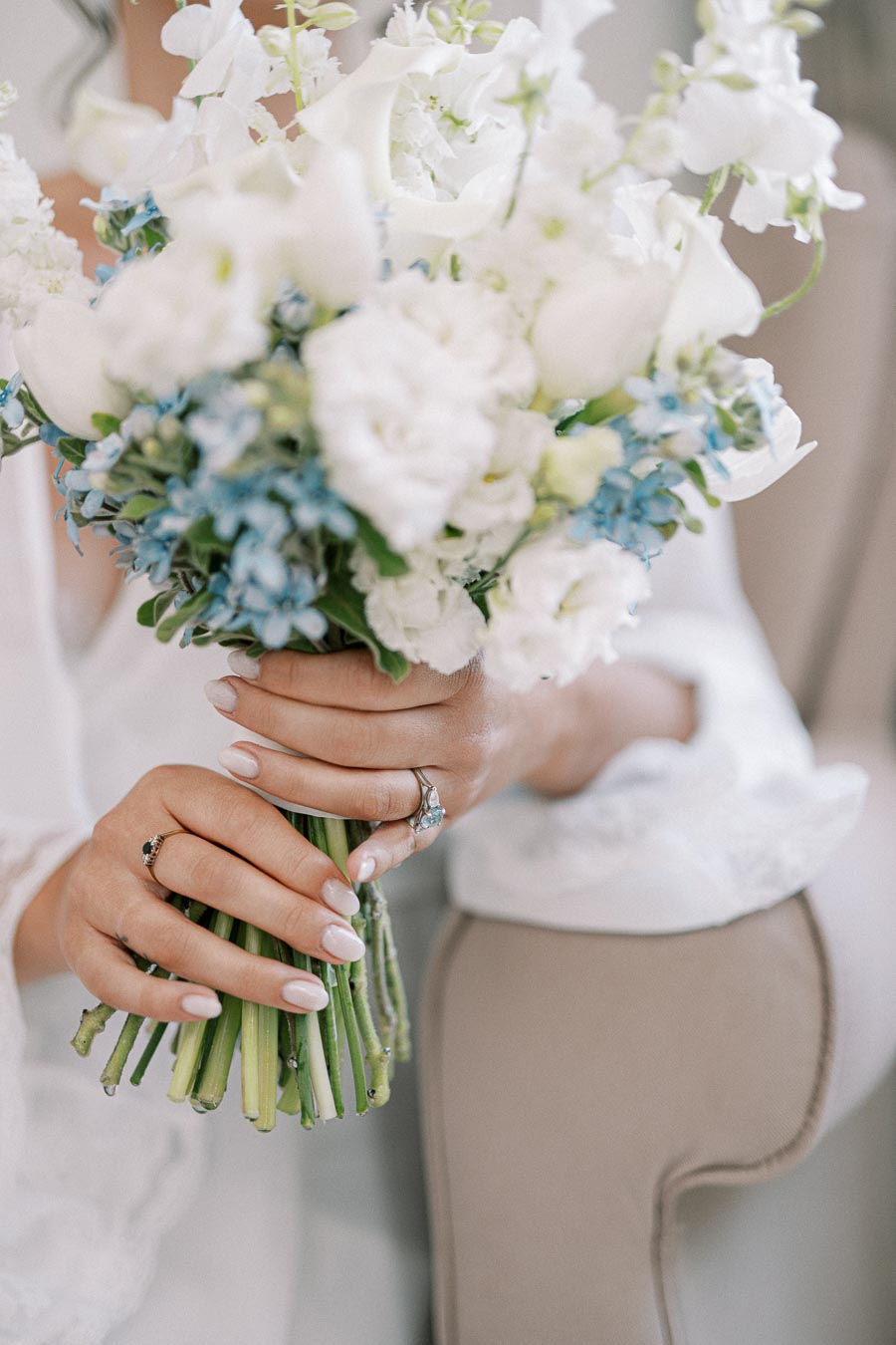 Close-up of a bride holding an elegant bouquet of white and blue flowers, showcasing her delicate manicure and diamond rings.