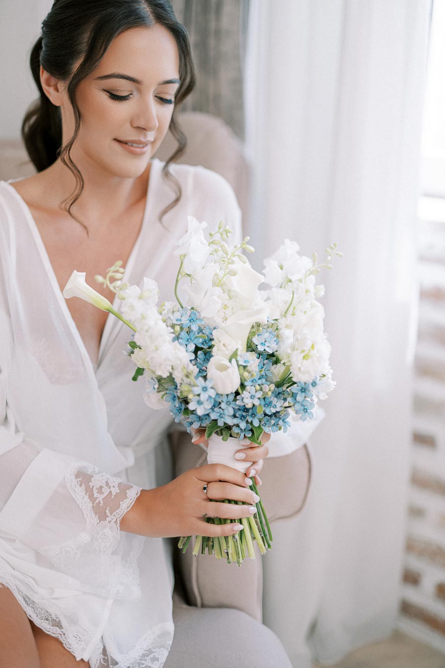 Young woman in a bridal robe holding a bouquet of white and blue flowers, preparing for her wedding day indoors.