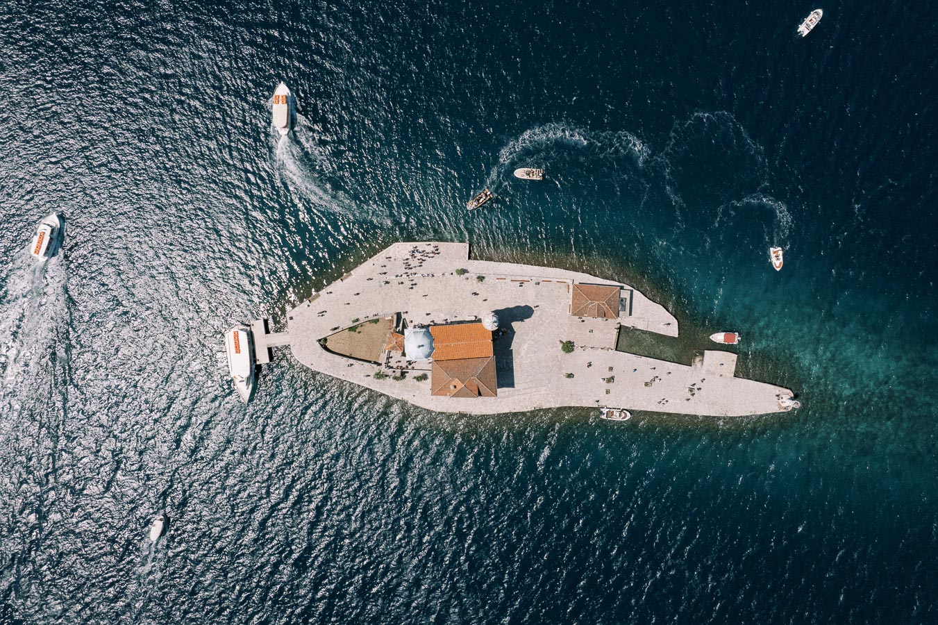 Aerial view of a small island with a church and buildings surrounded by deep blue ocean waters, with several boats navigating around the island.