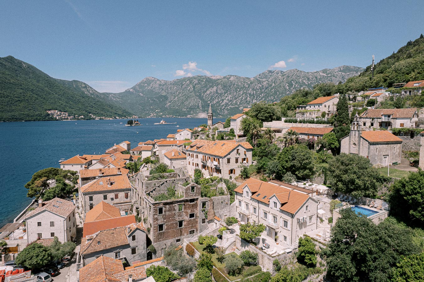 Scenic view of a coastal Mediterranean village with historic stone houses and terracotta roofs, nestled by the azure waters and surrounded by lush green hills and distant mountains under a clear blue sky.