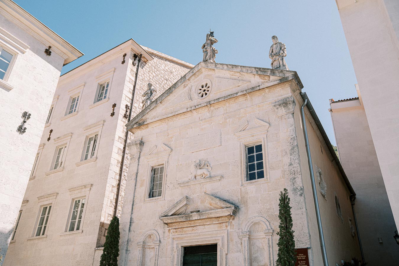 Historic stone church facade with statues on the roof, set against a clear blue sky, surrounded by neighboring buildings.
