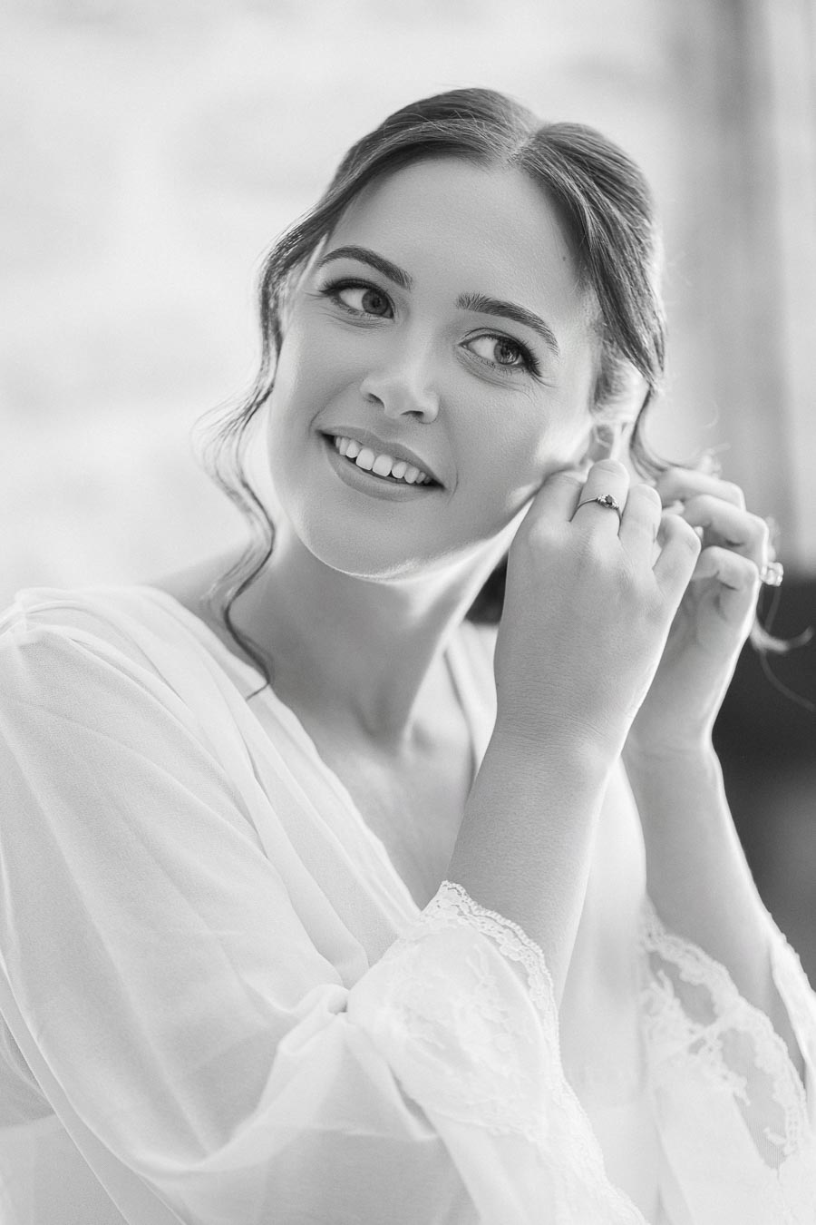 A bride getting ready for her wedding, smiling while putting on earrings, captured in a black-and-white portrait.