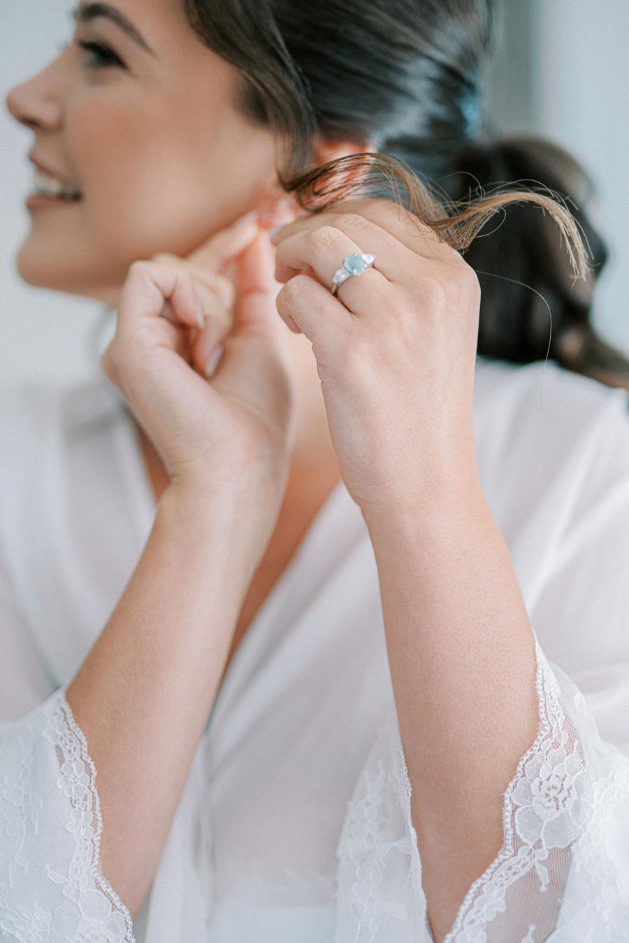 Young woman in a white lace robe adjusting her earring, close-up view of her face and hand with an engagement ring.