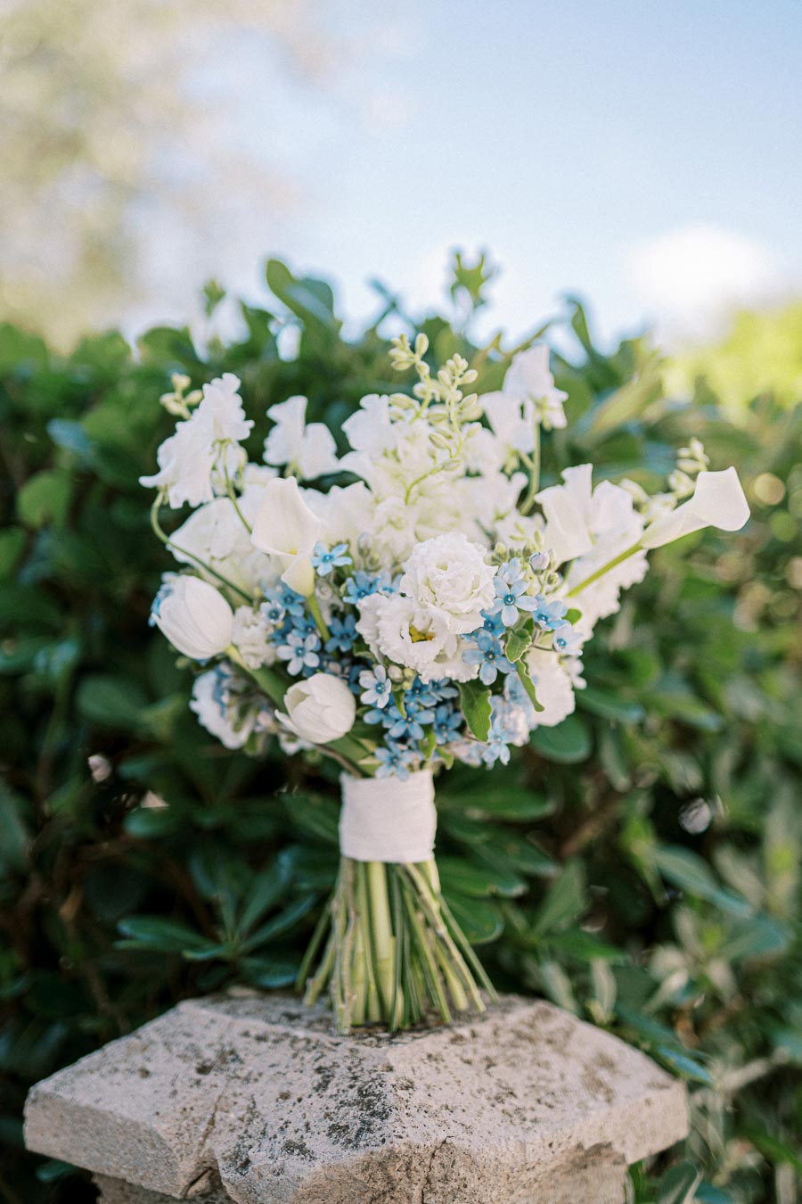 Bouquet of white and blue flowers with greenery, placed on a stone ledge outside against a blurred green foliage background.