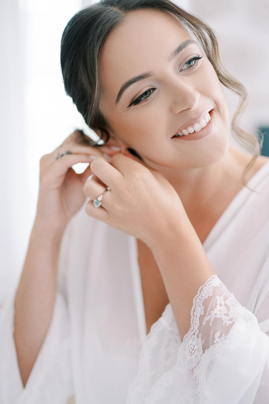 A bride getting ready, smiling as she adjusts her earring while wearing a white, lace-trimmed robe.