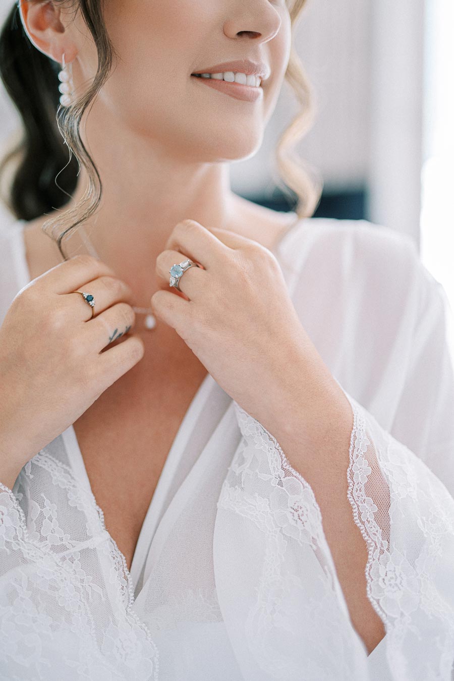 Woman in a white lace robe with elegant jewelry, focusing on her hands with diamond and gemstone rings and a pearl necklace.