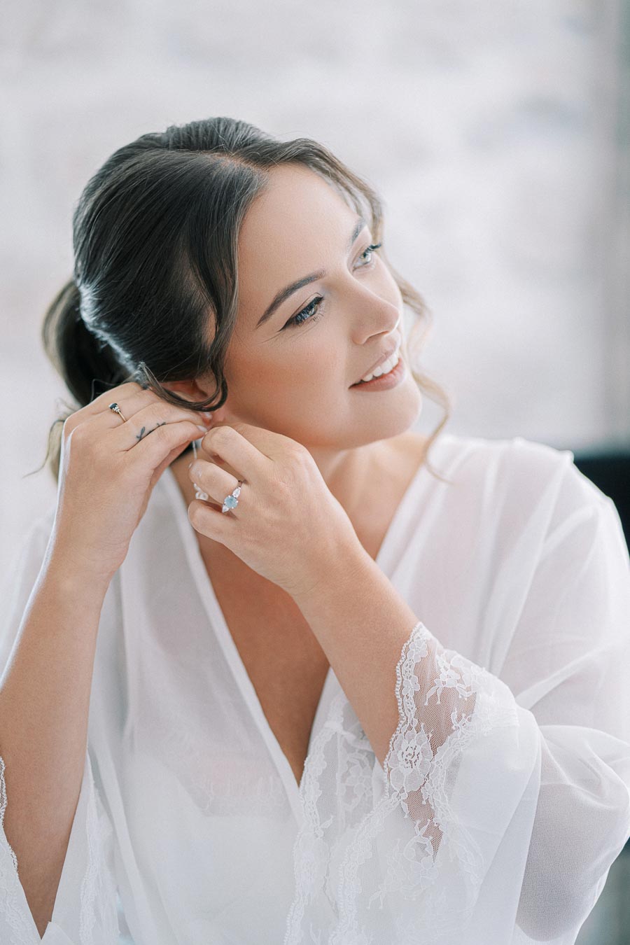 Bride getting ready on wedding day, wearing a white lace robe and putting on earrings.