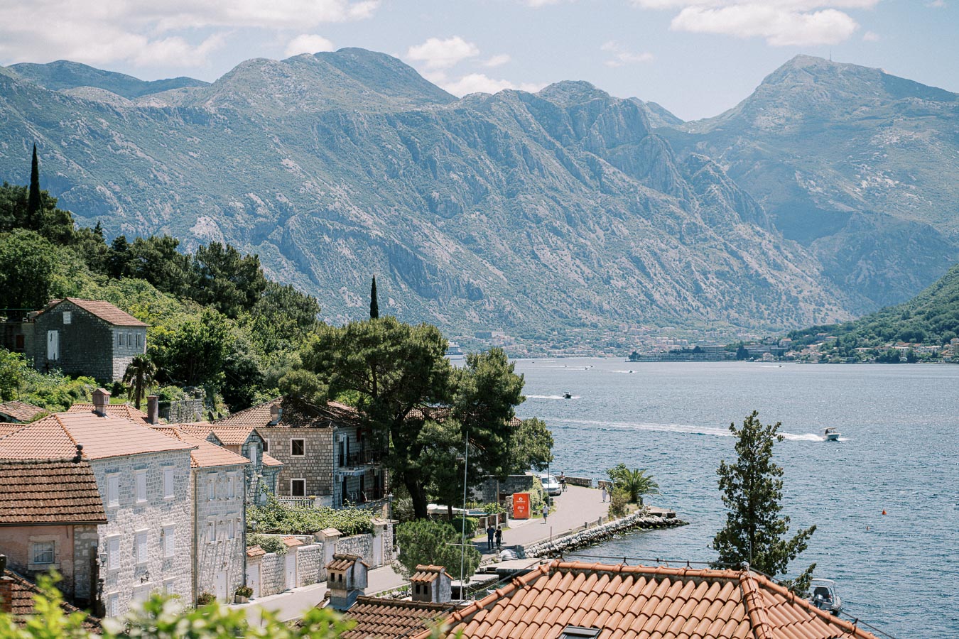 Scenic view of coastal village with terracotta-roofed houses and lush greenery, set against a backdrop of majestic mountains and a tranquil bay with boats, under a clear blue sky.