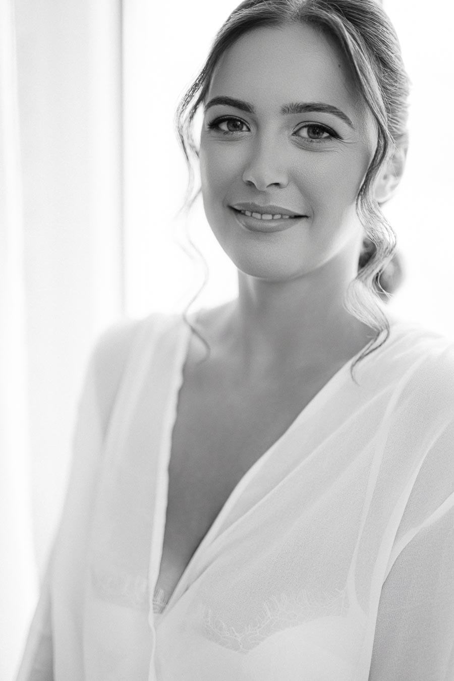 Black and white portrait of a smiling woman with wavy hair, wearing a sheer blouse, standing by a softly lit window.