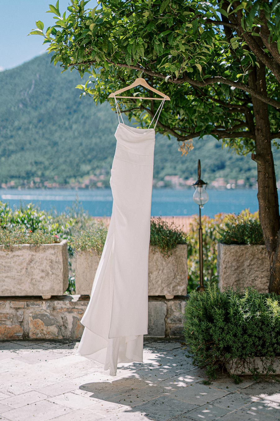 Elegant white wedding dress hanging from a tree branch in a picturesque outdoor setting with a view of a lake and mountains in the background.