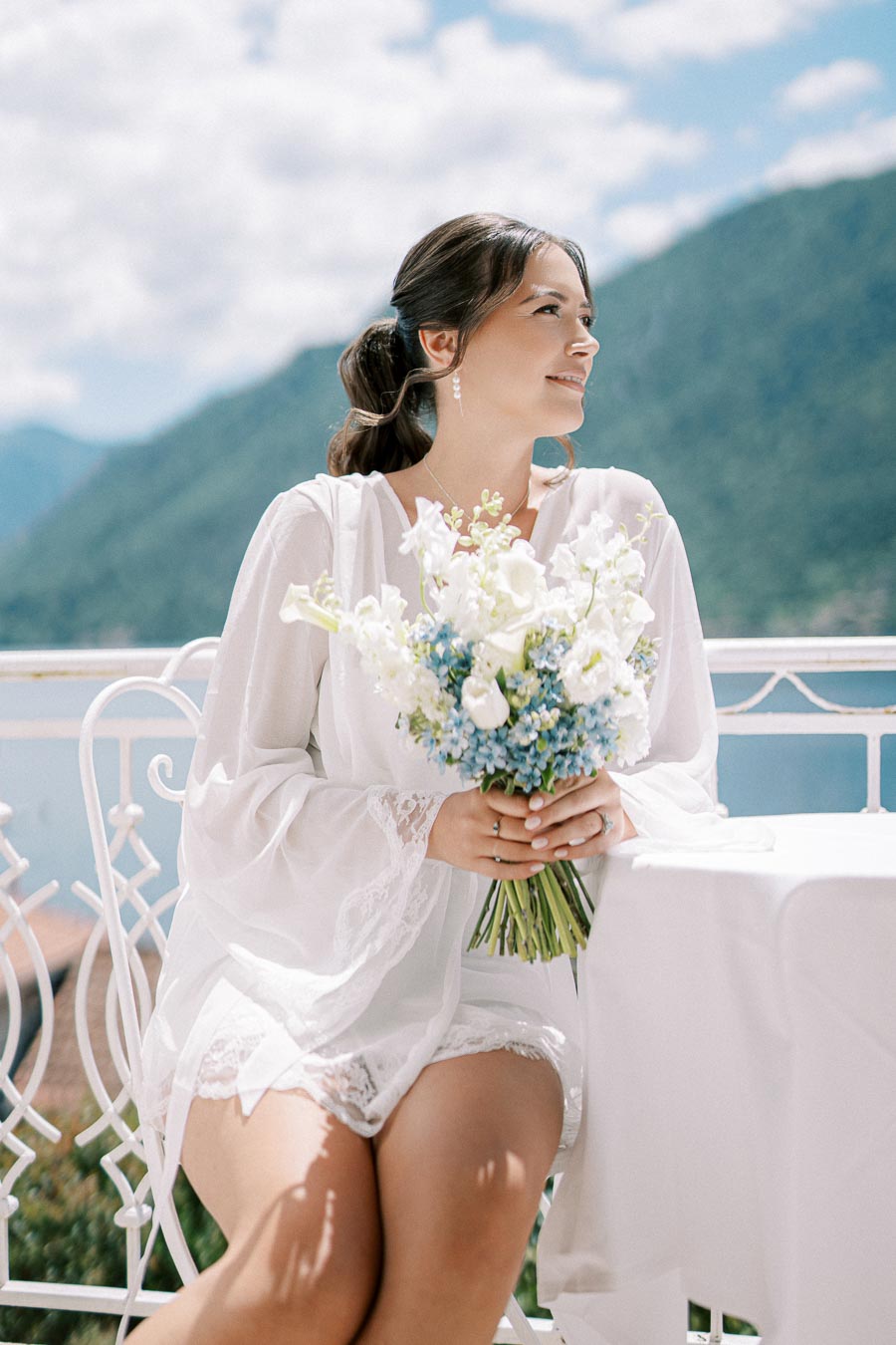 Young woman in a white dress holding a bouquet of white and blue flowers, seated on a balcony with a scenic mountain and lake view in the background.