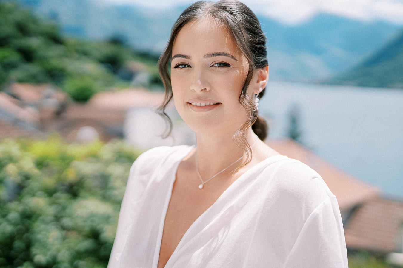 Smiling woman in a white dress with an elegant hairstyle, standing outdoors with a scenic view of a lake and mountains in the background