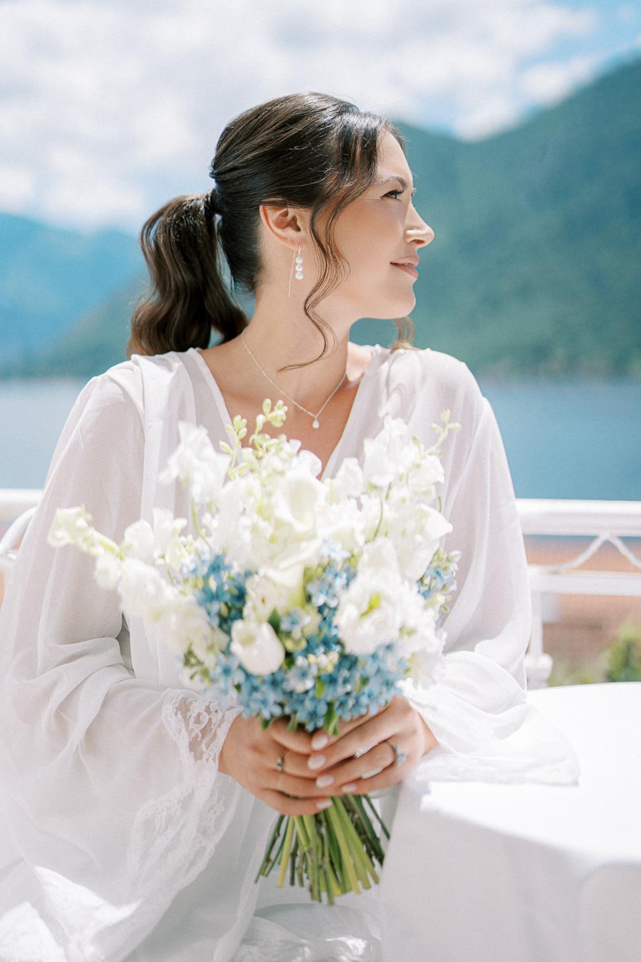 Elegant bride holding a bouquet of white and blue flowers with a scenic mountain lake backdrop