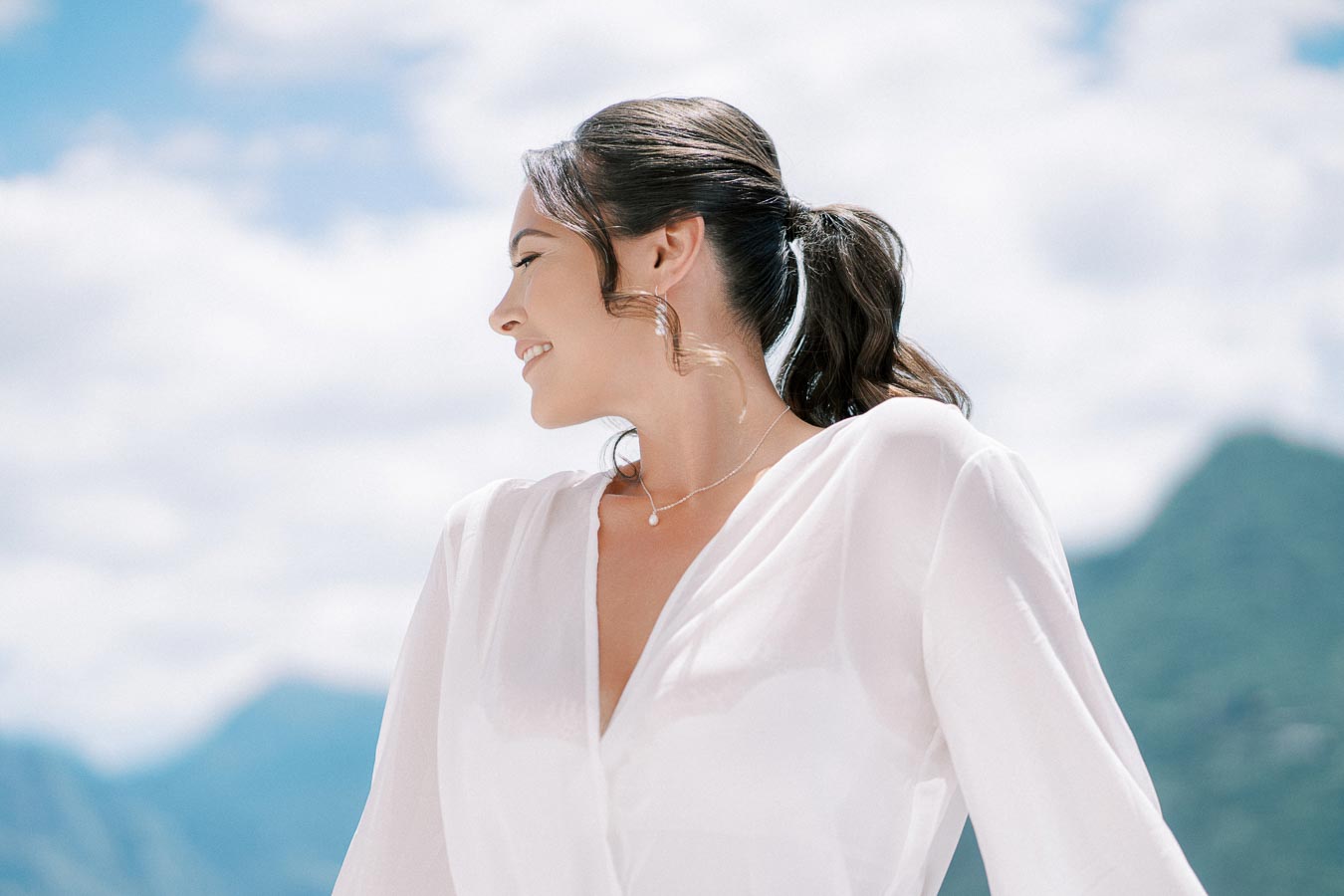 Young woman smiling and looking to the side, wearing a white blouse, with a mountainous landscape and blue sky in the background.