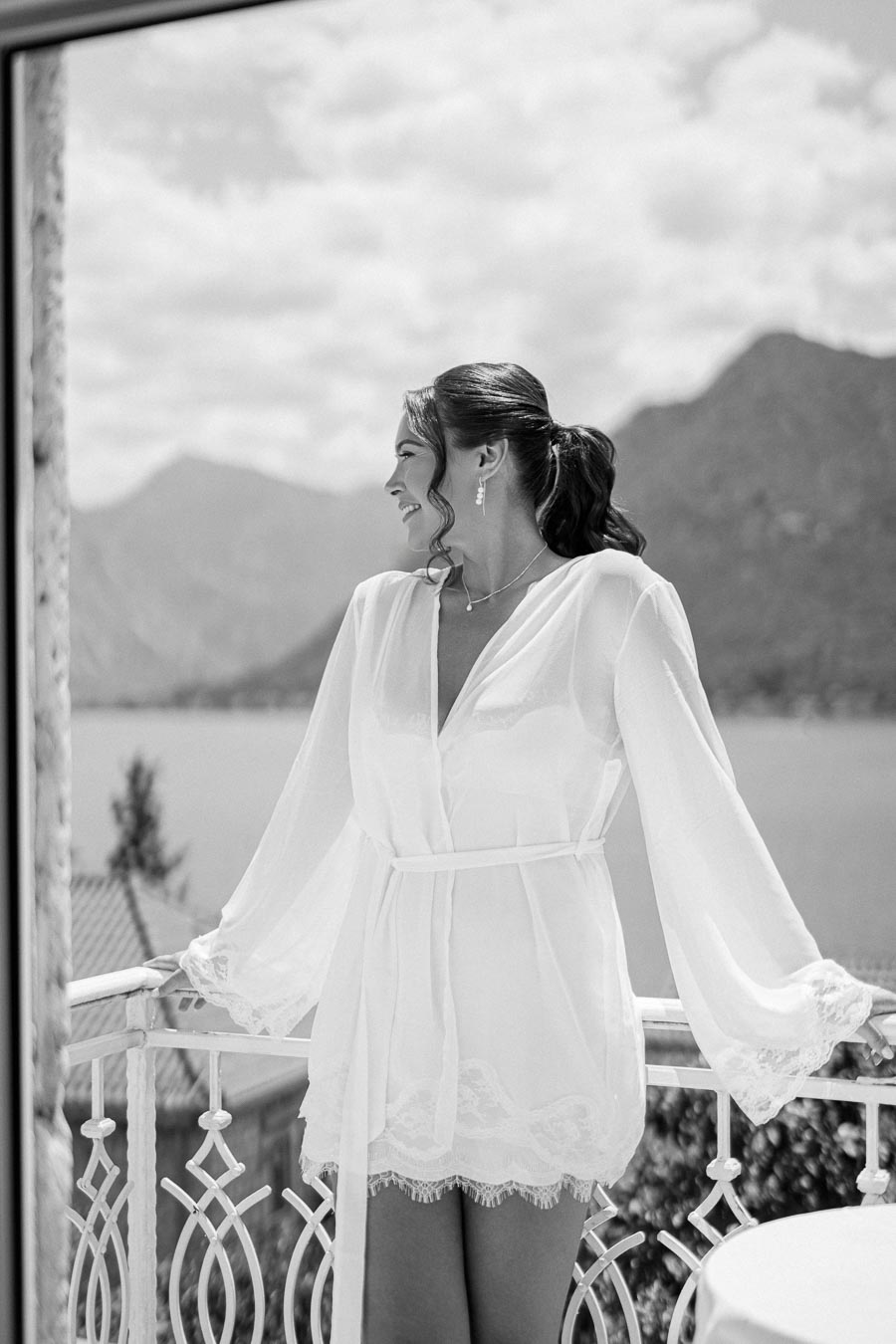 Black and white photo of a woman in a white robe on a balcony overlooking a scenic lake and mountains, exuding elegance and serenity.