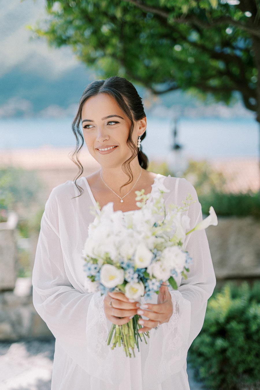 A smiling bride in a white dress holding a bouquet of white and blue flowers, standing outdoors with a scenic background.