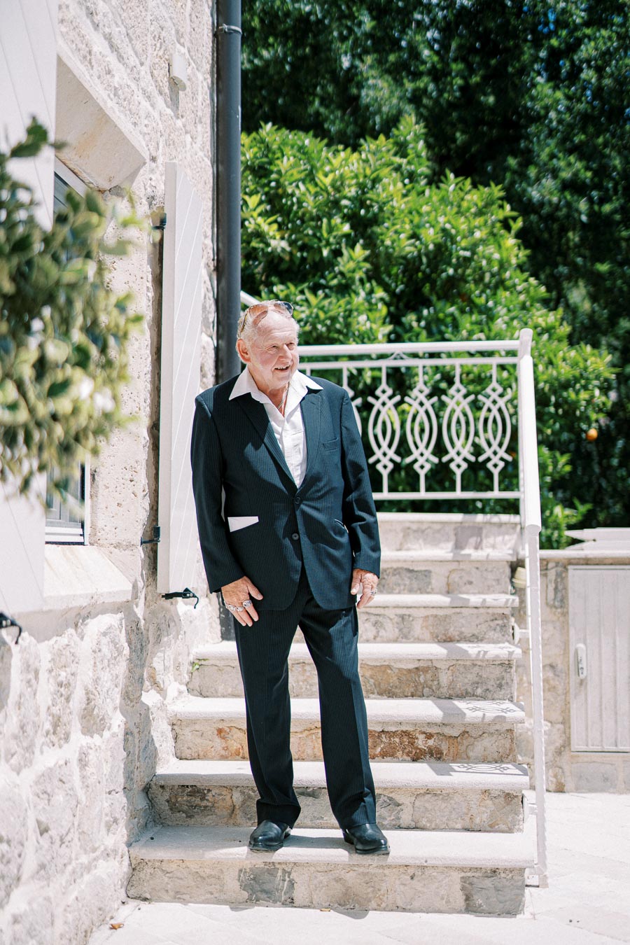 Senior man in a formal suit standing on stone steps next to a historic building with green foliage in the background.
