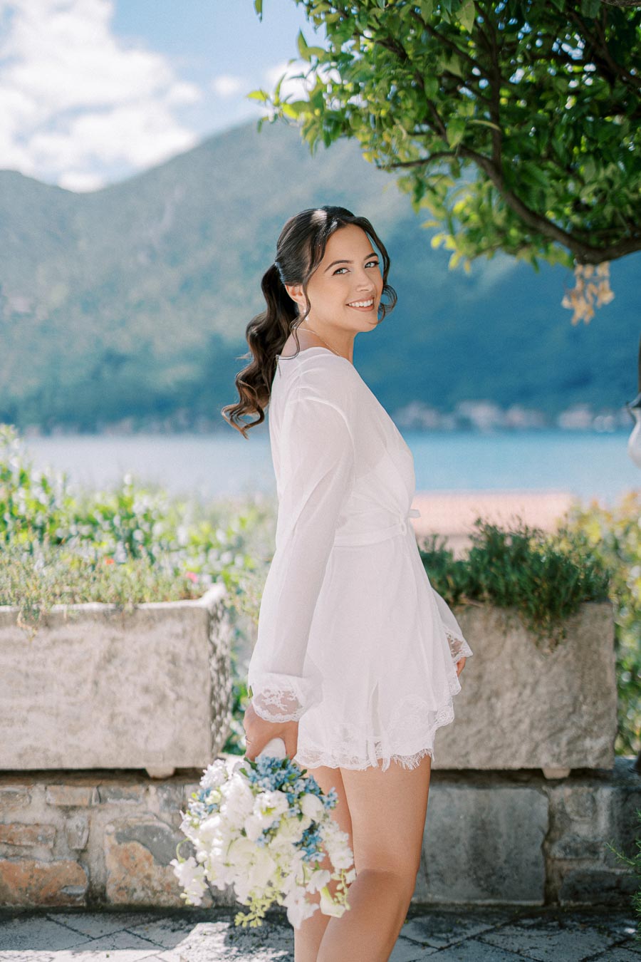 A woman in a white dress holding a bouquet of flowers, smiling with a mountainous landscape and a lake in the background on a sunny day.