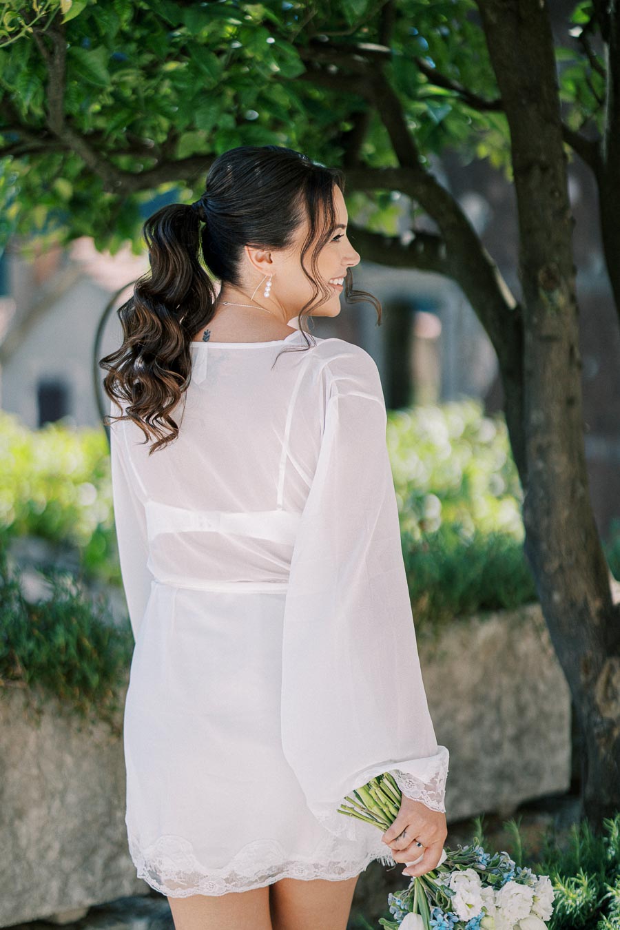 Young woman in elegant white robe and ponytail, holding a bouquet of flowers, smiling under a lush green tree in a garden setting.
