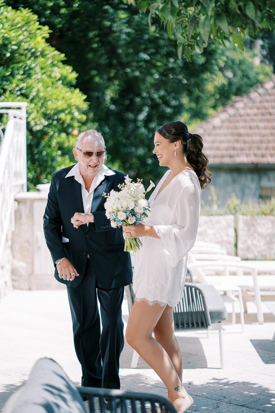 Smiling bride holding a bouquet walking arm-in-arm with an older man in a suit outdoors, surrounded by lush greenery and rustic architecture.