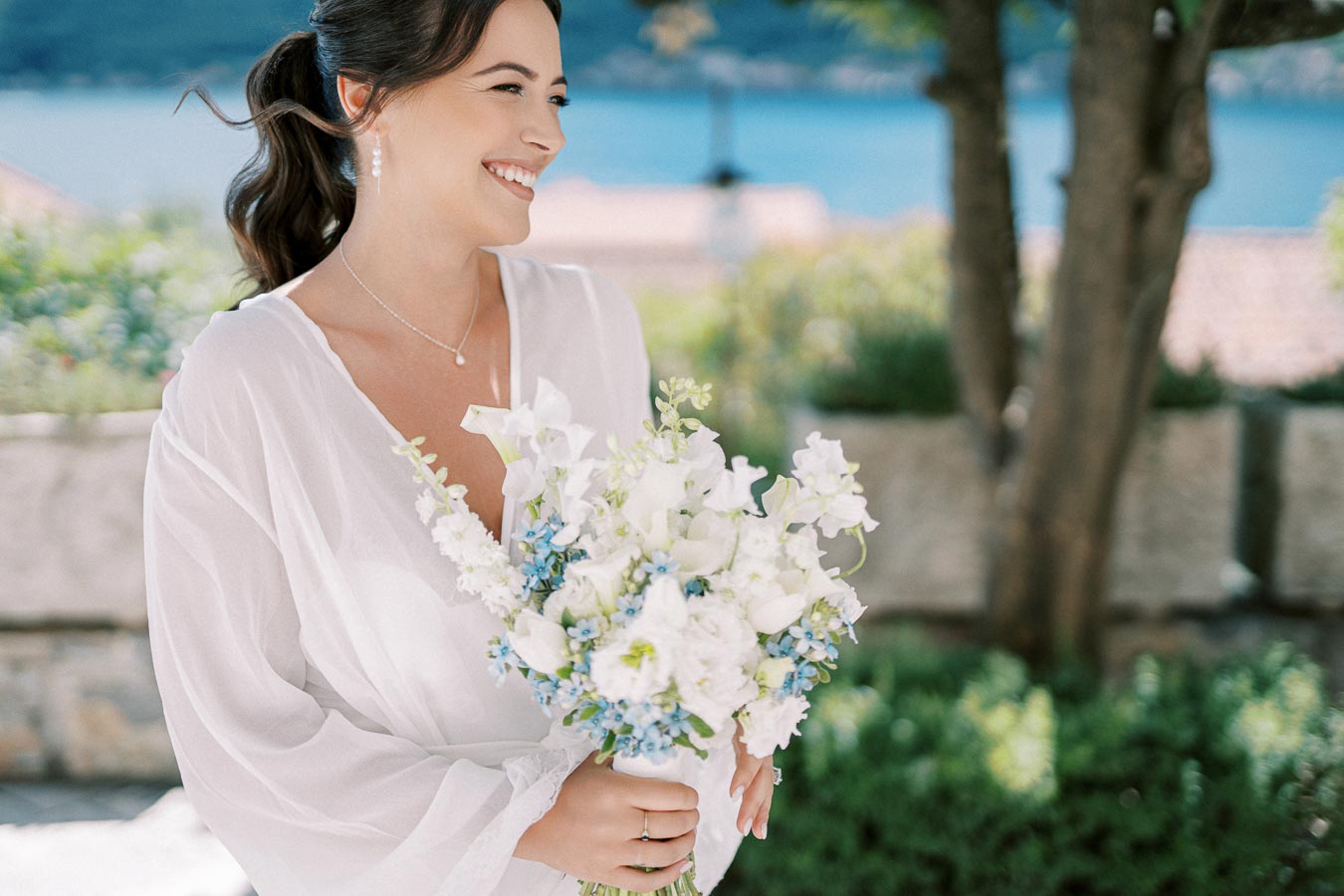 A smiling bride holding a bouquet of white and blue flowers, wearing a white dress, stands outdoors with a backdrop of greenery and a view of the water.