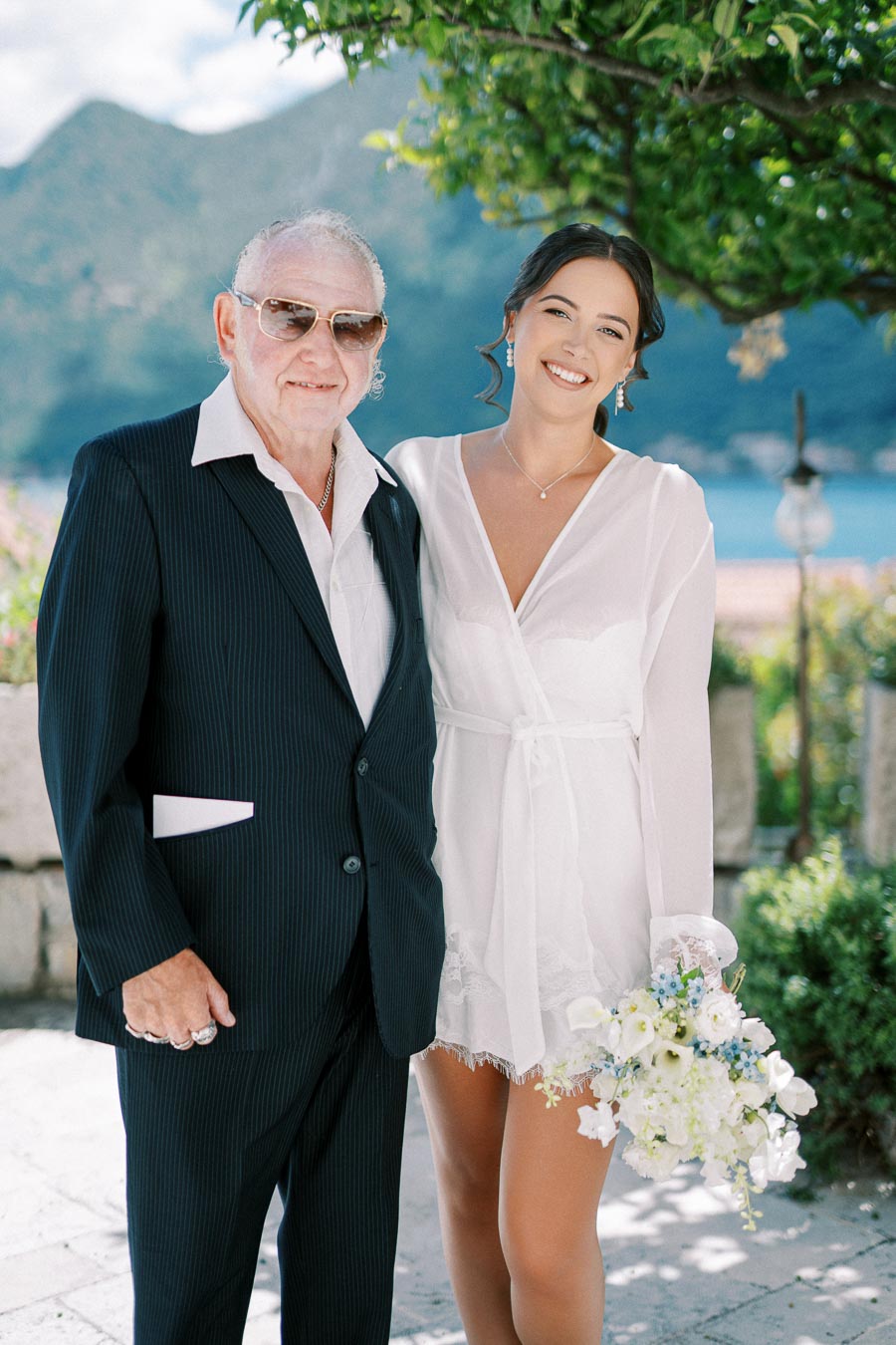 A smiling young woman in a white dress holds a bouquet of flowers while posing with an older man in a suit and sunglasses, in a picturesque outdoor setting with mountains in the background.