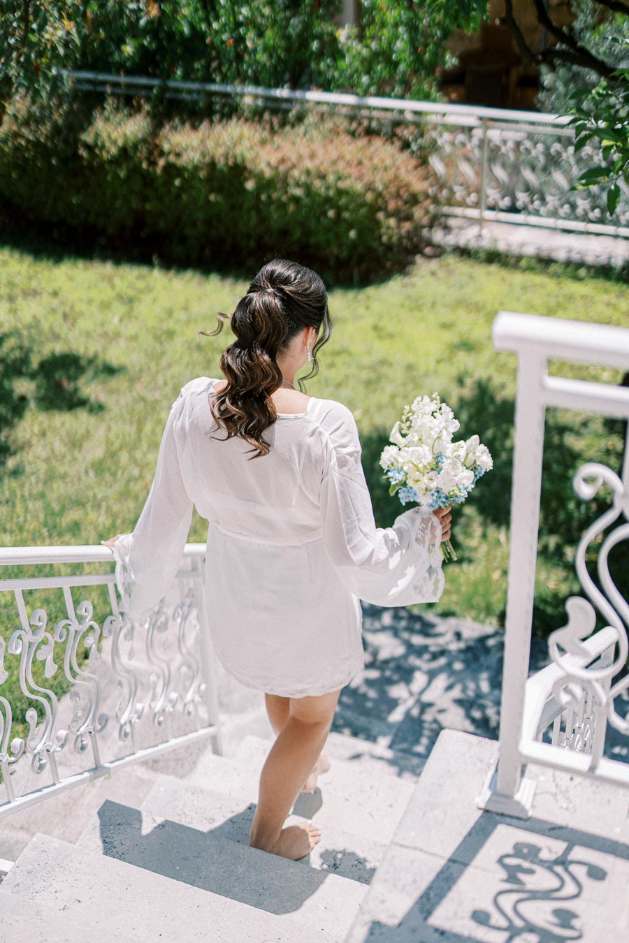 A woman in a white robe holding a bouquet of flowers descends a decorative outdoor staircase surrounded by greenery.