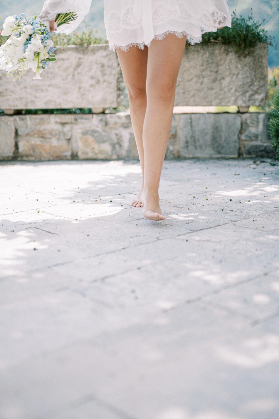 A barefoot bride walks gracefully on a stone path, holding a delicate bouquet of white and blue flowers, with sunlight casting soft shadows around her.