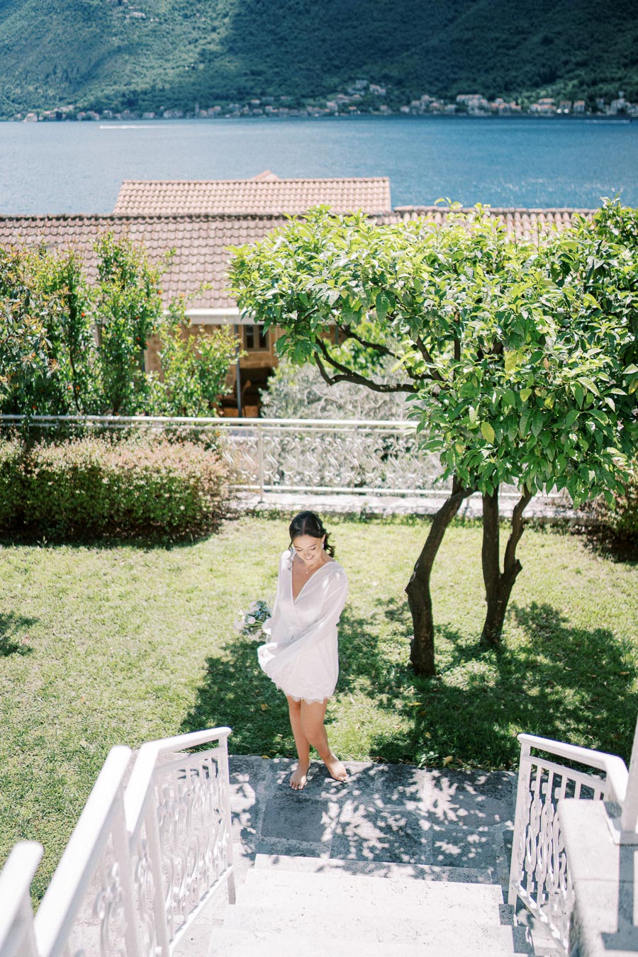 A woman in a white dress stands barefoot on a sunlit garden path, surrounded by lush greenery, with a scenic view of a lake and hills in the background.